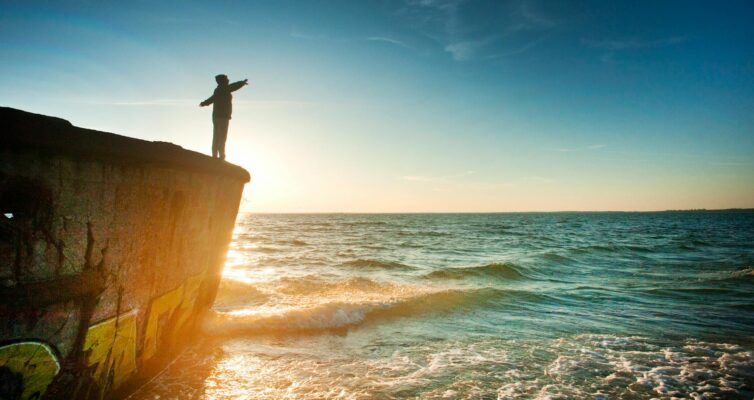 silhouette of person on cliff beside body of water during golden hour